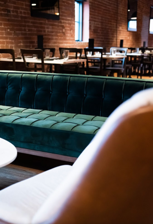 A cozy restaurant interior featuring a green velvet couch and wooden tables in a warm, brick-walled setting.