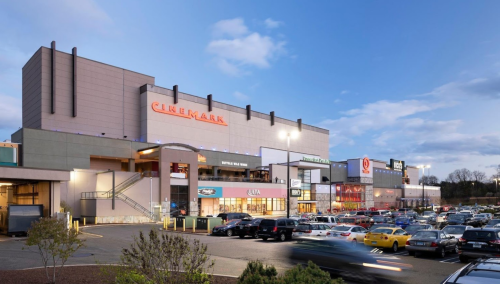 A shopping center with a Cinemark theater, featuring various stores and a parking lot filled with cars under a blue sky.