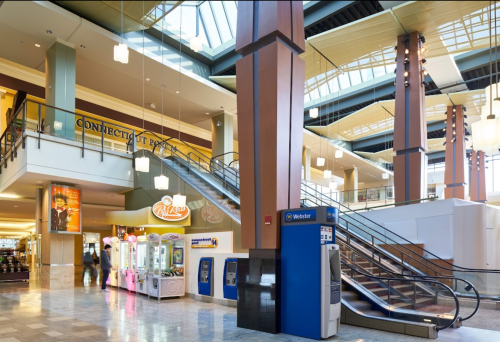Interior view of a shopping mall featuring escalators, kiosks, and a food court area with bright lighting.