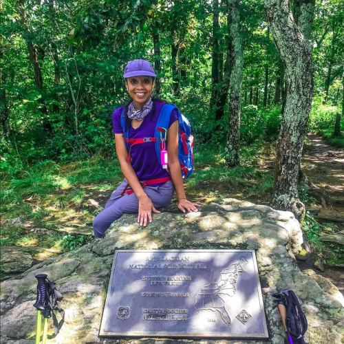 A hiker poses beside a trail marker in a lush green forest, wearing a backpack and a hat.