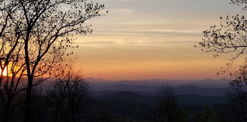 A serene sunset over rolling mountains, framed by silhouetted trees against a colorful sky.
