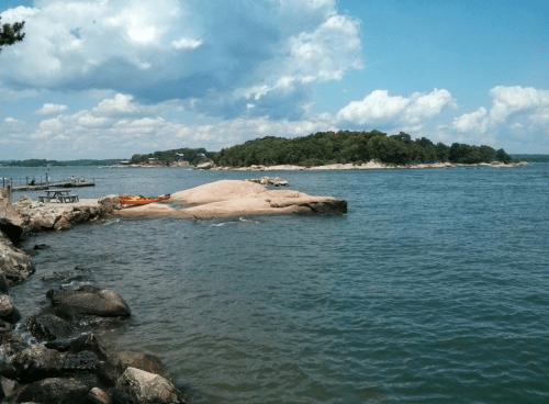A rocky shoreline with calm water, an island in the distance, and a partly cloudy sky.