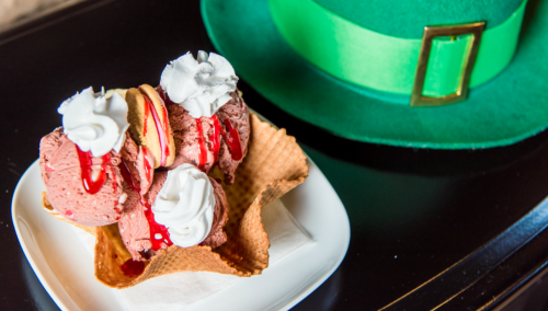A dessert bowl with three scoops of pink ice cream topped with whipped cream and syrup, next to a green leprechaun hat.