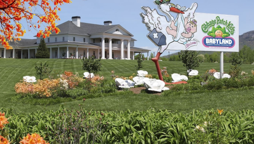 A large house with columns in the background, featuring a colorful sign for Cabbage Patch Kids Babyland and blooming flowers.