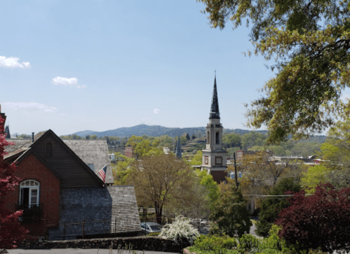 A scenic view of a town with a church steeple, trees, and mountains under a clear blue sky.