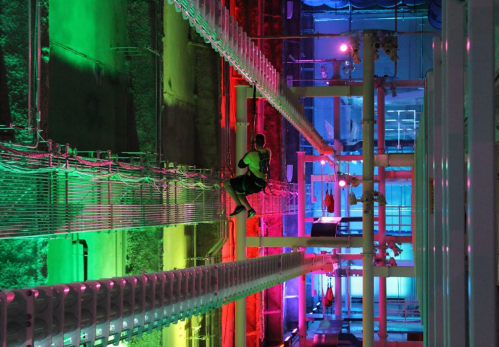 A person in a neon-lit industrial space, suspended on a rope against colorful walls.