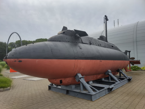 A black and red submarine displayed outdoors on a concrete platform, surrounded by greenery and modern buildings.