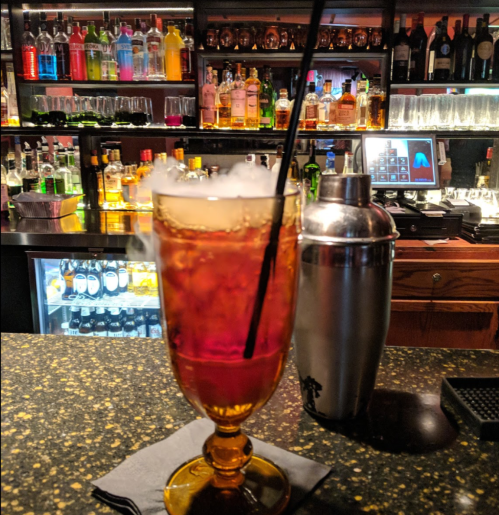 A tall cocktail glass filled with a dark drink and smoke, set on a bar with various bottles in the background.