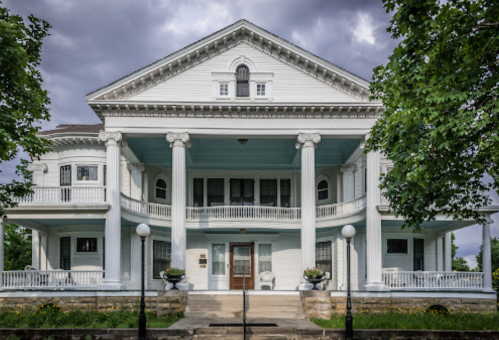 A large, elegant white mansion with columns, a balcony, and a landscaped front yard under a cloudy sky.