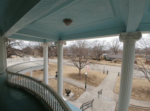 View from a porch with decorative columns, overlooking a tree-lined street and a grassy area.