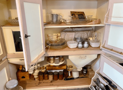 A cluttered kitchen cabinet filled with various glassware, jars, and utensils, showcasing a mix of vintage and modern items.