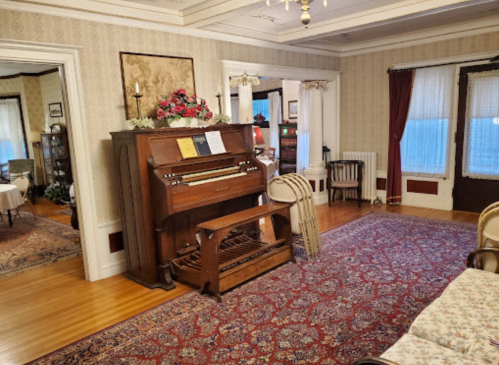 A vintage piano in a cozy, elegantly decorated room with patterned wallpaper and a large area rug.