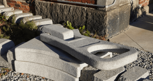 A large, gray stone sculpture resembling a stylized tool, resting on a gravel surface near a brick wall.