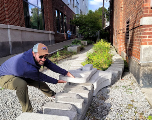A man in a cap is placing stones along a curved path in a landscaped area beside a building.
