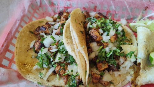 Two tacos filled with grilled chicken, chopped onions, and cilantro, served in a red and white basket with lime wedges.