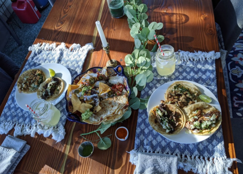 A wooden table set with tacos, nachos, and drinks, surrounded by greenery and a candle.