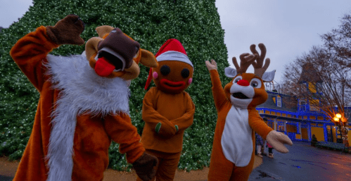 Three festive mascots in holiday costumes pose in front of a large Christmas tree, smiling and waving.