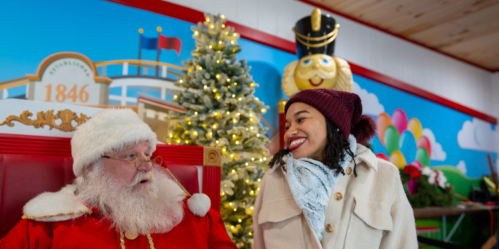 A joyful woman smiles at Santa Claus in a festive setting with a Christmas tree and colorful decorations.