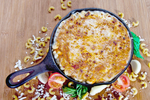 A baked macaroni and cheese dish in a cast iron skillet, surrounded by pasta, tomatoes, and fresh herbs on a wooden surface.