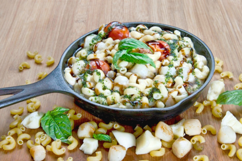 A cast-iron skillet filled with pasta, cherry tomatoes, and basil, surrounded by scattered pasta on a wooden surface.