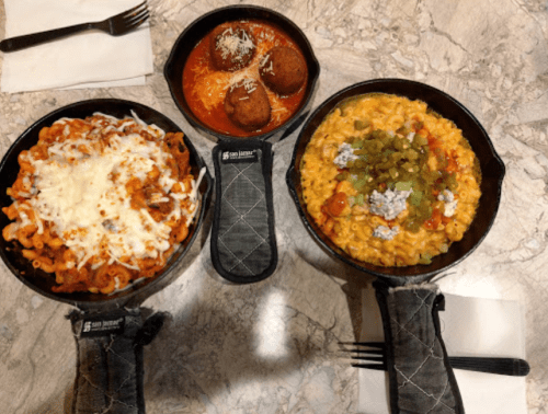 Three cast iron dishes of pasta: one with cheese, one with meatballs in sauce, and one with macaroni and green toppings.
