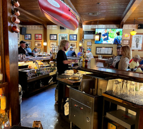 A cozy bar scene with patrons, a bartender serving drinks, and a TV showing a nature program in a rustic setting.
