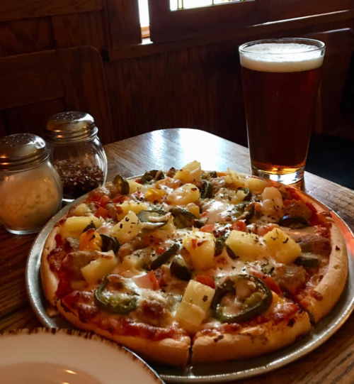 A pizza topped with pineapple, jalapeños, and tomatoes, served with a pint of beer and condiments on a wooden table.