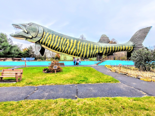 A large, colorful fish sculpture stands in a grassy area with visitors nearby, under a cloudy sky.