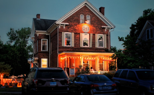 A charming brick house with warm lights, surrounded by parked cars, during twilight.