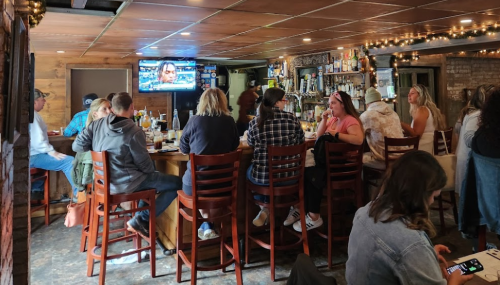 A lively bar scene with patrons seated at a wooden counter, watching a TV and enjoying drinks in a cozy atmosphere.