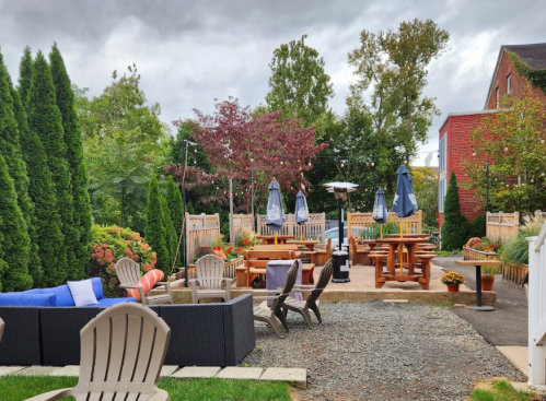 Outdoor seating area with wooden tables, umbrellas, and lush greenery under a cloudy sky. Cozy and inviting atmosphere.