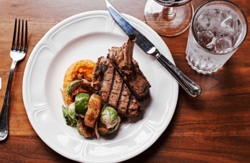Grilled lamb chop on a plate with Brussels sprouts and creamy polenta, accompanied by a glass of water and cutlery.