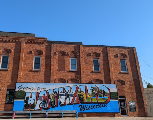 A colorful mural reading "Greetings from Hayward Wisconsin" on a brick building under a clear blue sky.