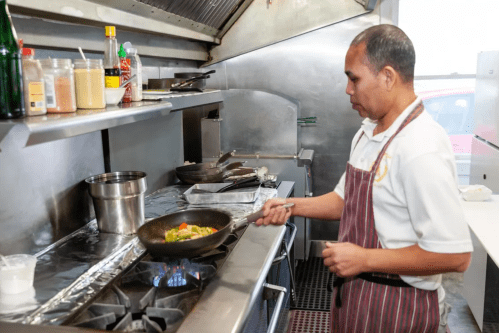 A chef in an apron cooks vegetables in a skillet over a gas stove in a commercial kitchen.