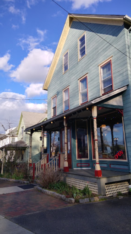 A blue, two-story building with a porch and an "Open" sign, surrounded by greenery and a cloudy sky.