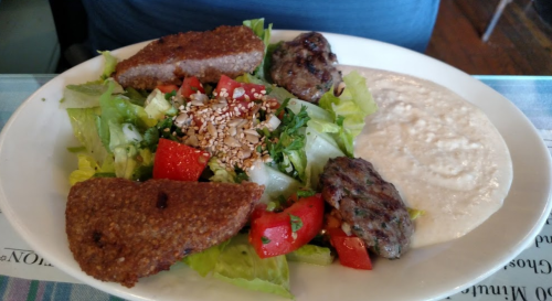 A plate with a salad topped with tomatoes and herbs, alongside two types of meat patties and a side of creamy sauce.