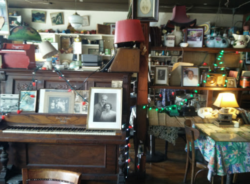 A cozy, vintage café interior featuring a piano, old photographs, and eclectic decor with festive lights.