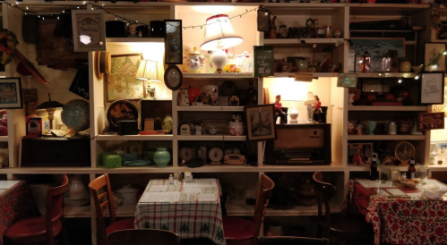 Cozy restaurant interior with vintage decor, shelves filled with antiques, and tables set with checkered tablecloths.