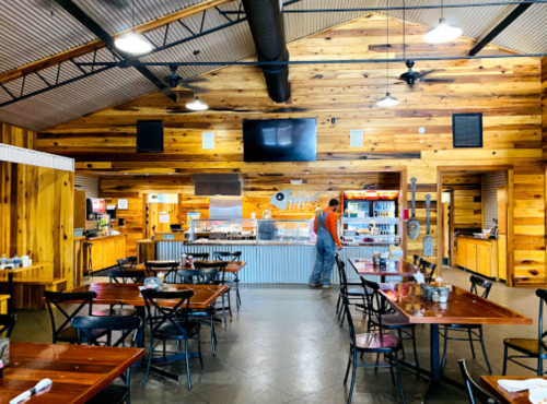 Interior of a rustic restaurant with wooden walls, tables, and a counter, featuring a staff member preparing food.