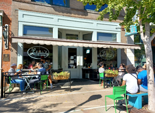 A bustling café with outdoor seating, people dining, and greenery along the sidewalk on a sunny day.