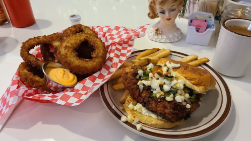 A plate with a fried chicken sandwich topped with veggies, served with fries, and a basket of onion rings on the side.