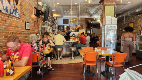 A bustling restaurant with brick walls, patrons dining at tables, and a bar area in the background.
