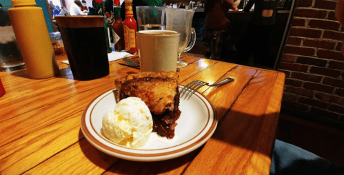 A slice of cherry pie with a scoop of vanilla ice cream, accompanied by a cup of coffee on a wooden table.