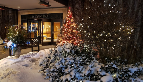 A snowy scene outside The Saint Paul hotel, featuring a decorated Christmas tree and twinkling lights on bushes.