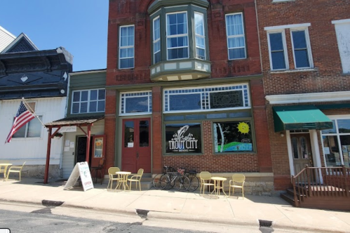 A charming storefront with "Trout City" sign, outdoor seating, and an American flag on a sunny day.