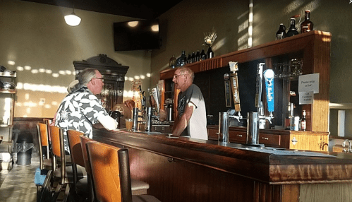 Two men converse at a bar with sunlight streaming through windows, surrounded by bottles and a cozy atmosphere.