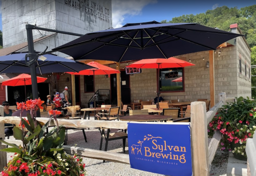 Outdoor seating area of Sylvan Brewing with umbrellas, flowers, and patrons enjoying the sunny day in Lanesboro, Minnesota.