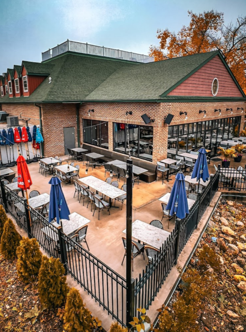 Outdoor dining area with tables, blue umbrellas, and a brick building in the background, surrounded by a black fence.