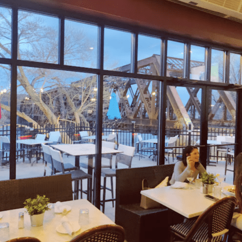 A cozy restaurant with large windows overlooking a bridge, featuring tables and a woman sitting at a table.