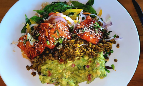 A colorful bowl featuring salmon, lentils, guacamole, and mixed vegetables, topped with sesame seeds.